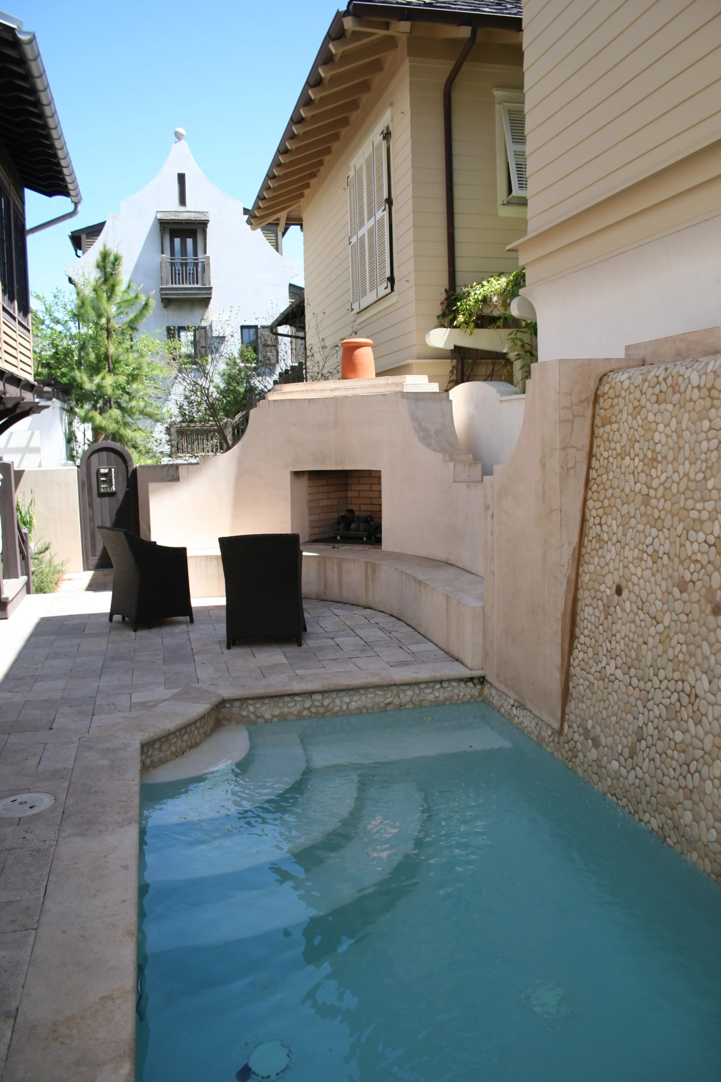 Private backyard featuring a small swimming pool with curved steps, two black outdoor chairs, a built-in fireplace, and nearby houses with balconies under a clear blue sky.
