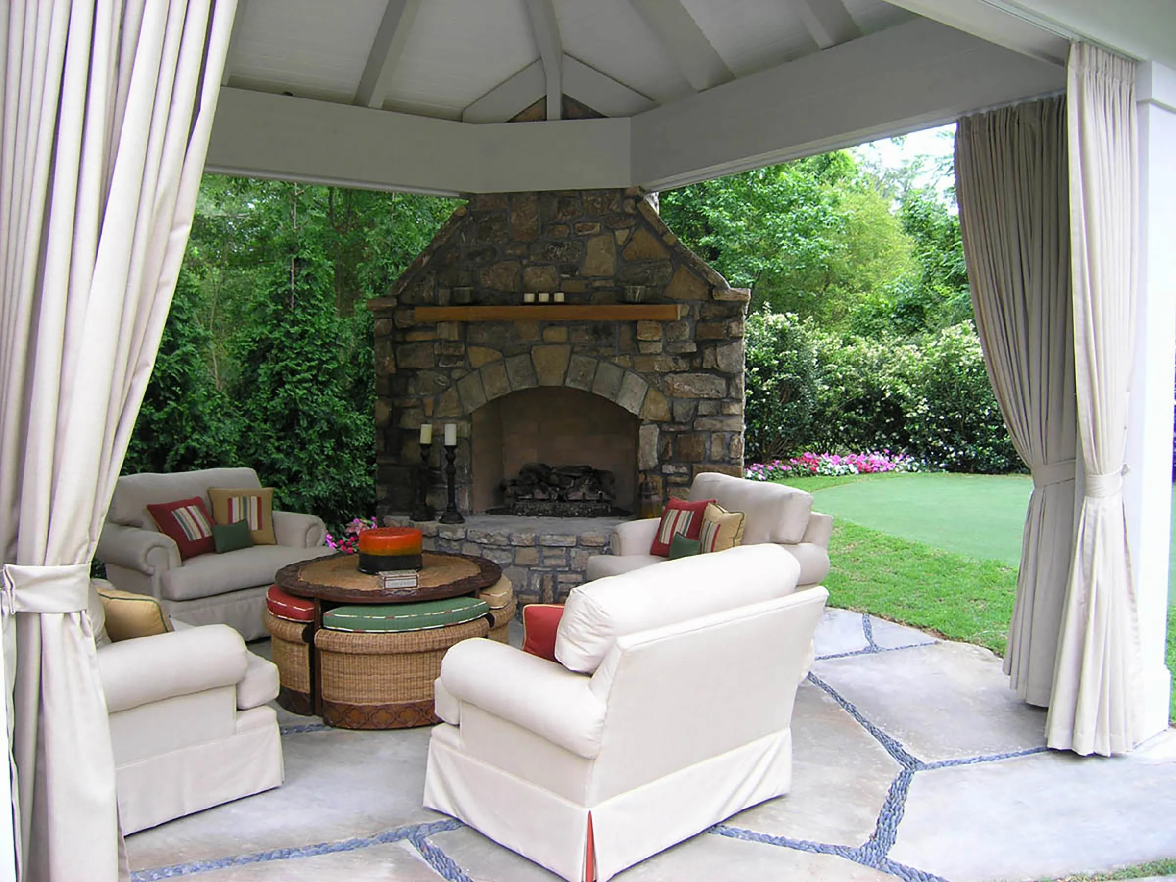 Outdoor living space with a stone fireplace, four white sofas, and colorful cushions, overlooking a garden with green grass and blooming flowers.