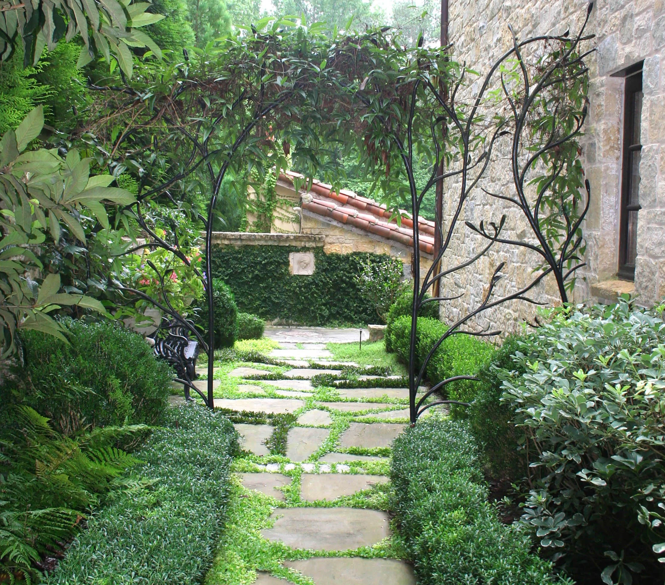 A lush garden with a stone pathway lined by green bushes and plants, an iron archway over the path, and a stone house wall on the right.