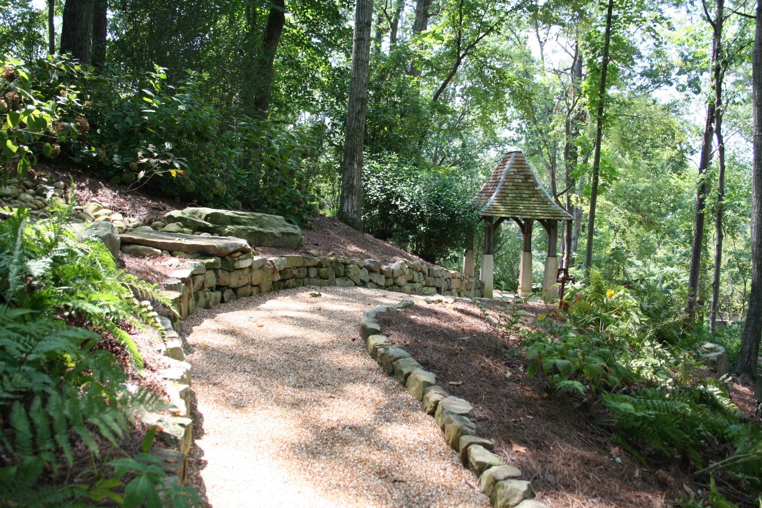 A gravel garden path winding through a lush, green forest with trees and ferns, leading to a small wooden gazebo with a shingled roof.