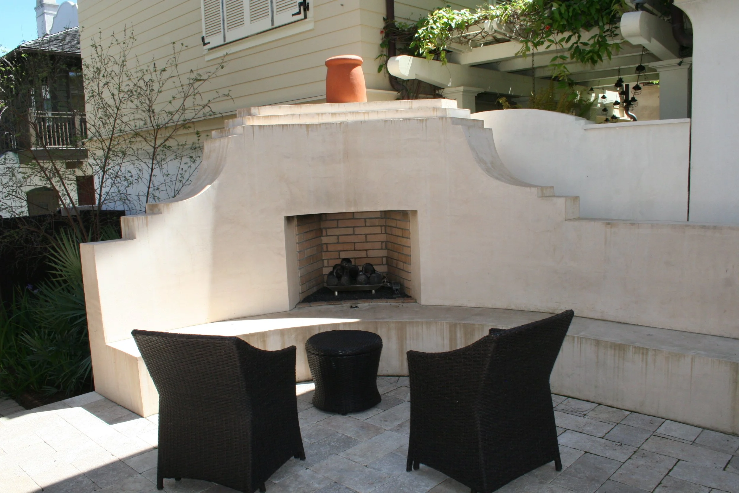 An outdoor patio area featuring a white concrete fireplace with a brick interior, two black wicker chairs, a black wicker side table, and some plants nearby.