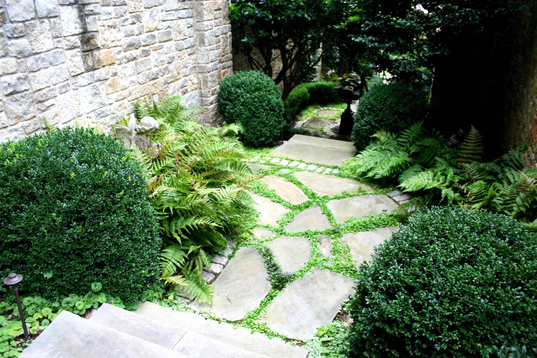 A lush garden pathway lined with trimmed bushes, ferns, and decorative statues, leading to a stone birdbath and a section of stone steps next to a brick wall.