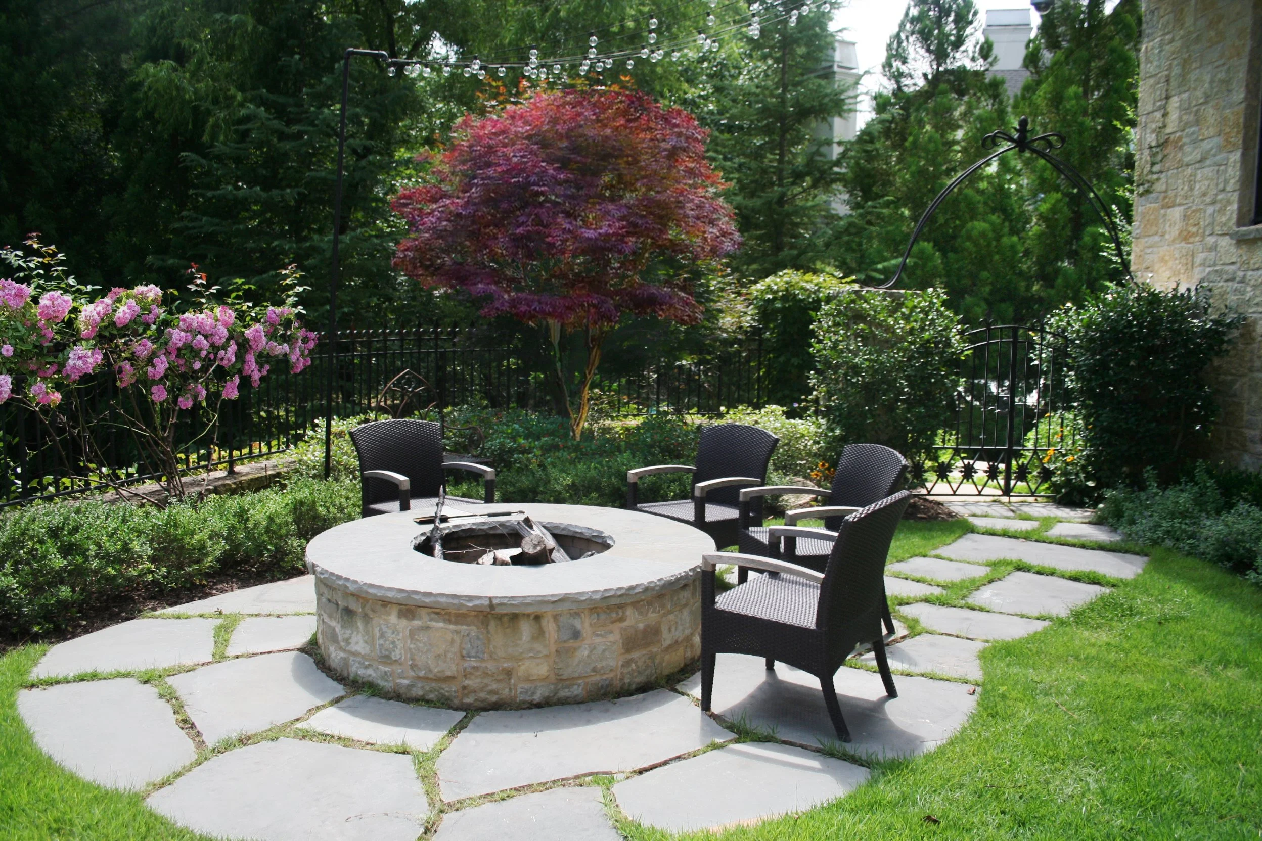 A backyard patio with a fire pit surrounded by four black chairs on a stone patio, lush green grass, and a garden with pink and purple flowering bushes and trees, enclosed by a black metal fence and archway.