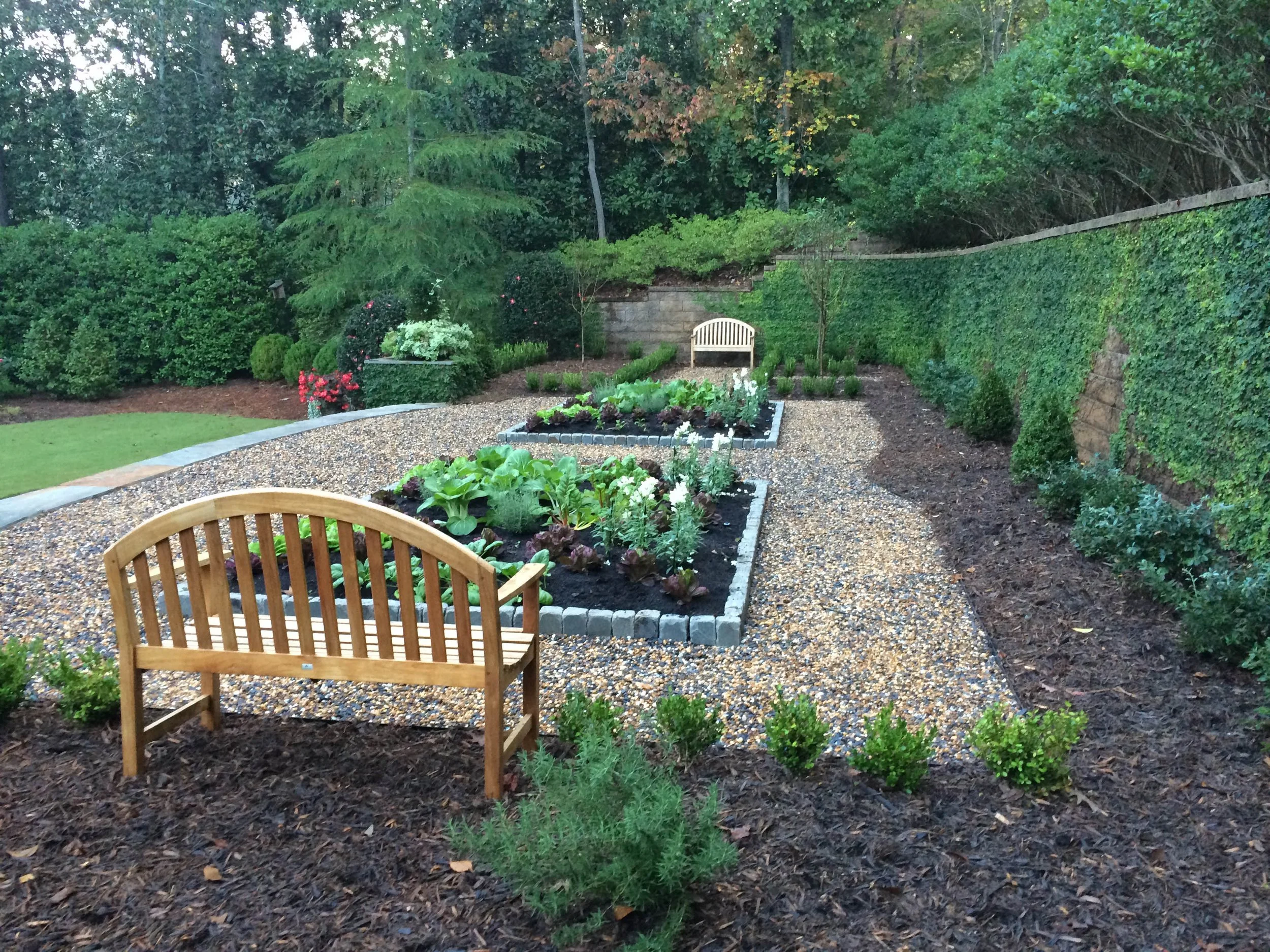 A landscaped backyard garden with a gravel pathway, raised garden beds with vegetables, a wooden bench, and lush trees and bushes in the background.