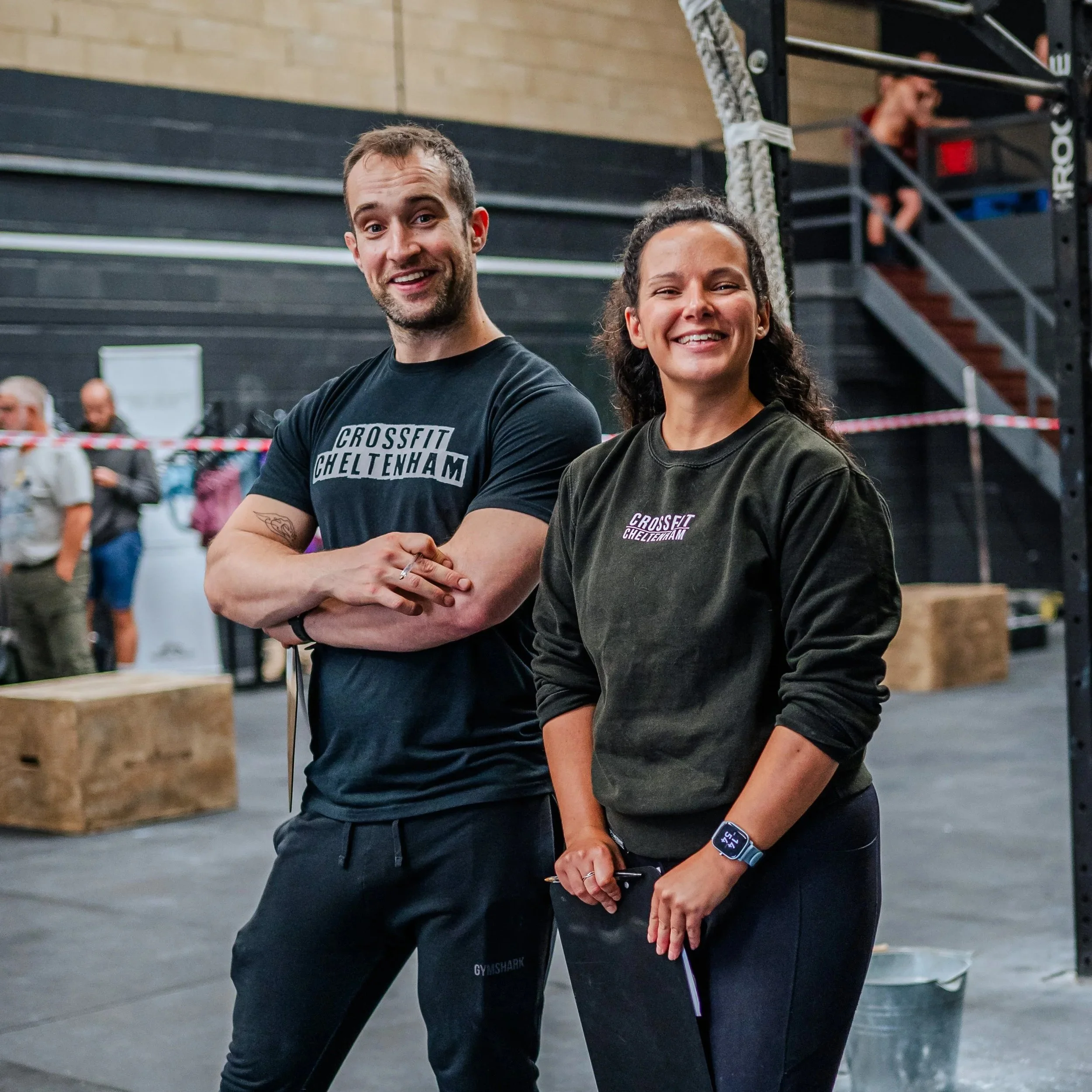 A man and a woman smiling at the camera inside a gym, wearing CrossFit Cheltenham shirts, with gym equipment and other people in the background.
