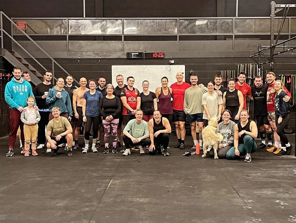Group photo of people and a dog in CrossFit Cheltenham, a gym, with some individuals wearing workout clothing, standing or kneeling in front of a whiteboard and gym equipment.