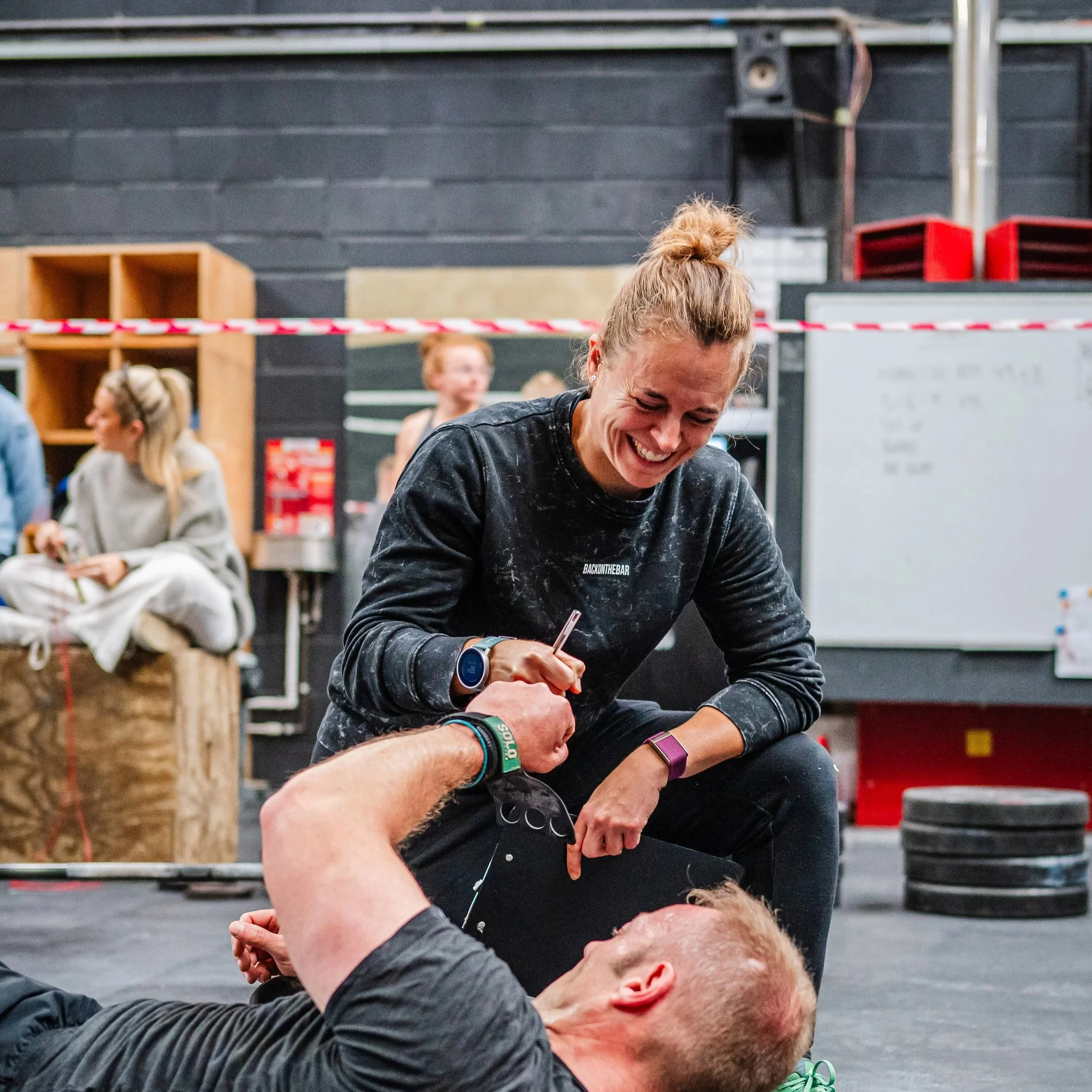 A woman has just judged a mans workout in a CrossFit competition in CrossFit Cheltenham, a gym. The man is lying on the floor and the lady is kneeling, they are laughing and sharing a moment. Other people are visible in the background.