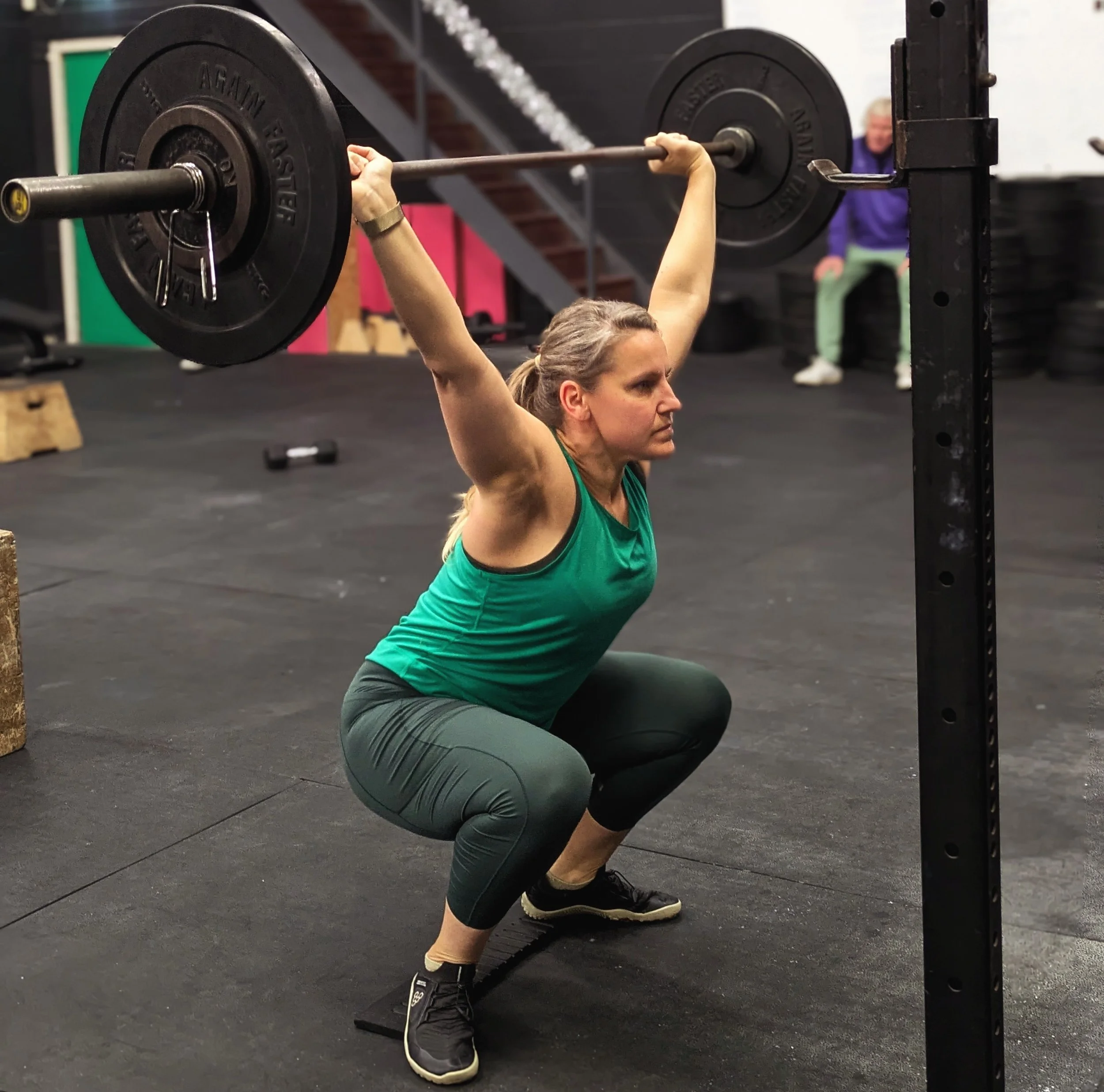 A woman wearing a green tank top and black leggings performing an overhead squat with a barbell in a CrossFit gym. She is squatting down with her arms extended overhead, holding the barbell. In the background, there is a person sitting on a bench.