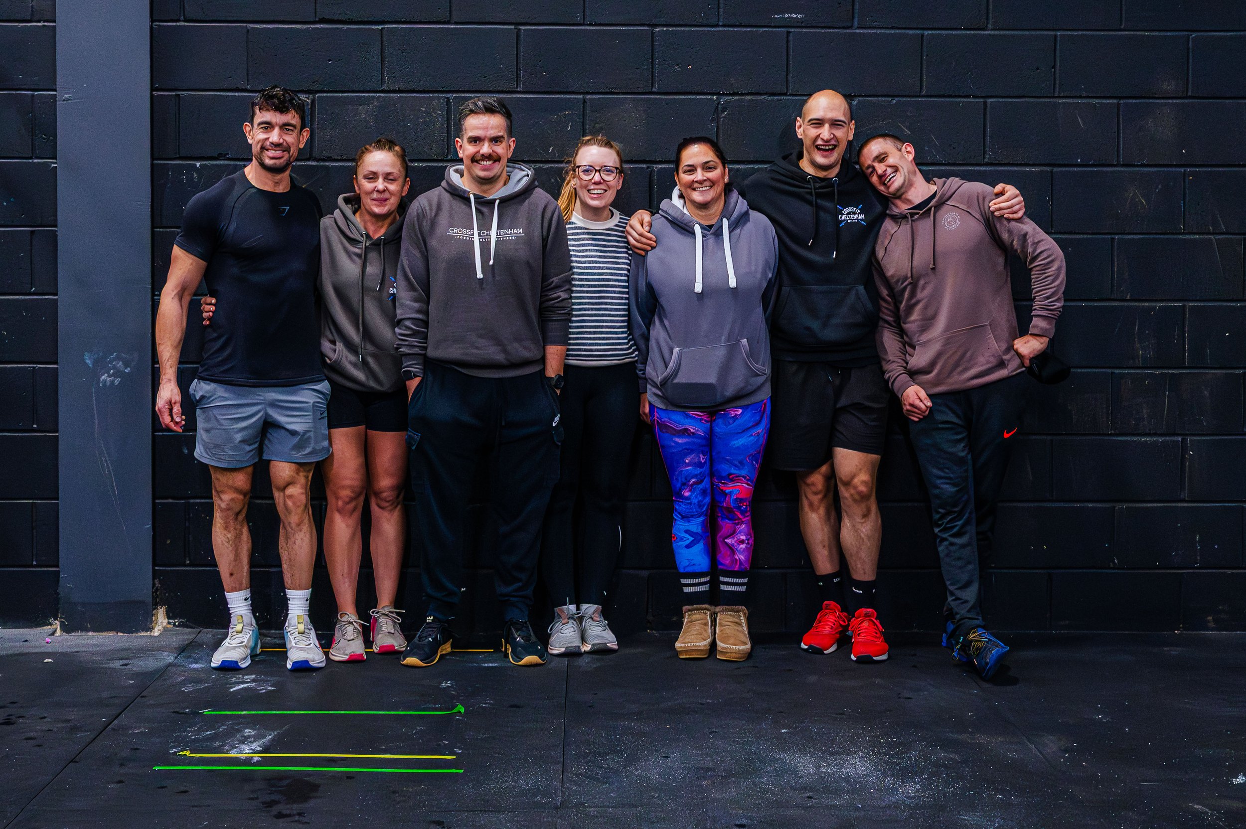 Group of seven people standing against a black wall, smiling and engaging in a fitness or workout session, with some wearing workout clothes and sneakers.