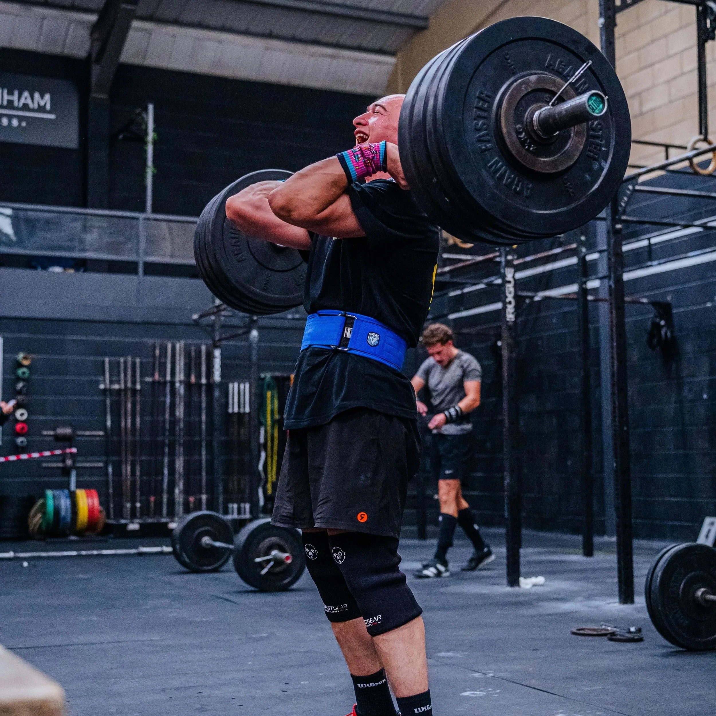 A man lifting a heavy barbell during a strength training workout in a gym, with another person working out in the background.