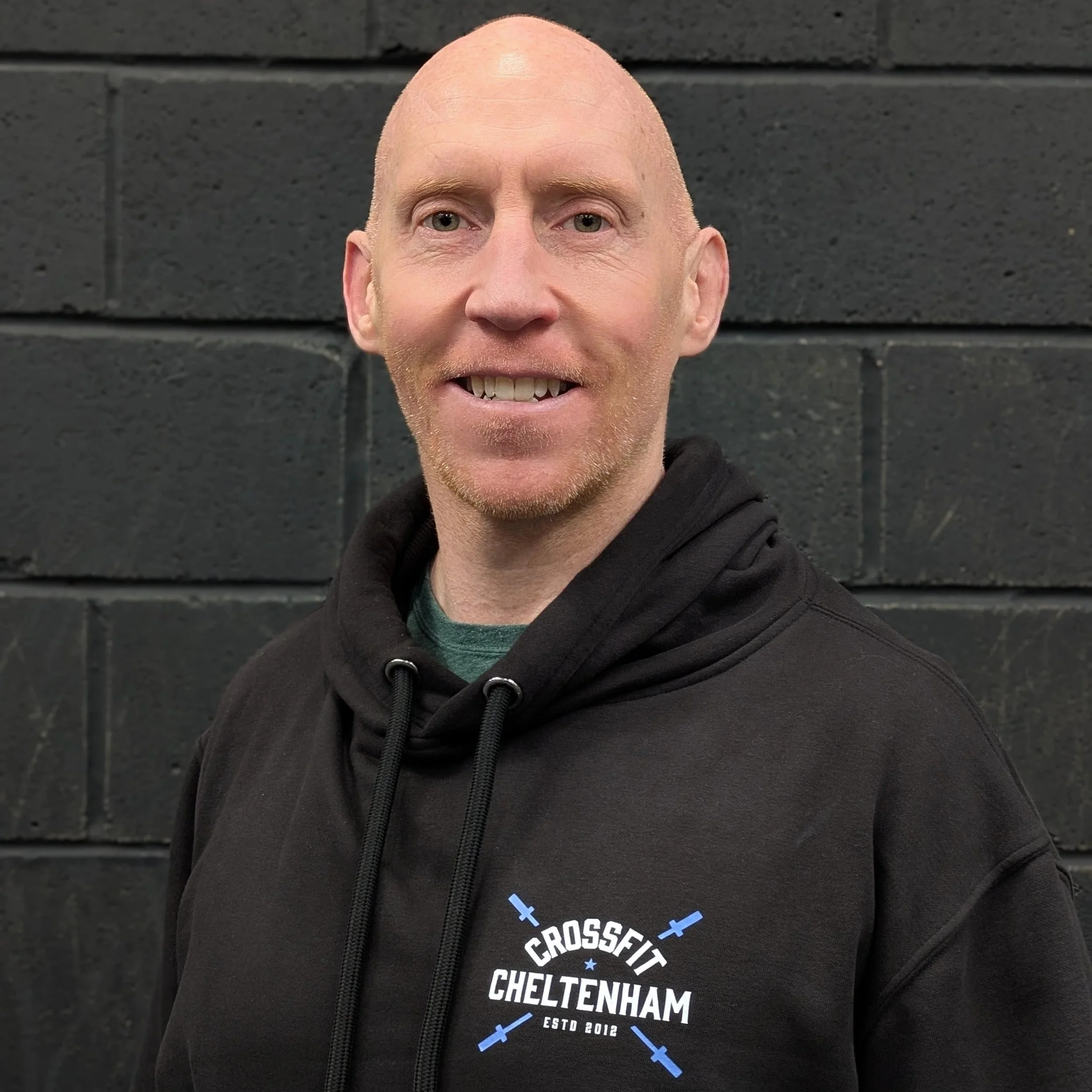 CrossFit Cheltenham head coach Steve Bunn is standing in front of a black brick wall, smiling, wearing a black hoodie with a CrossFit Cheltenham logo. 