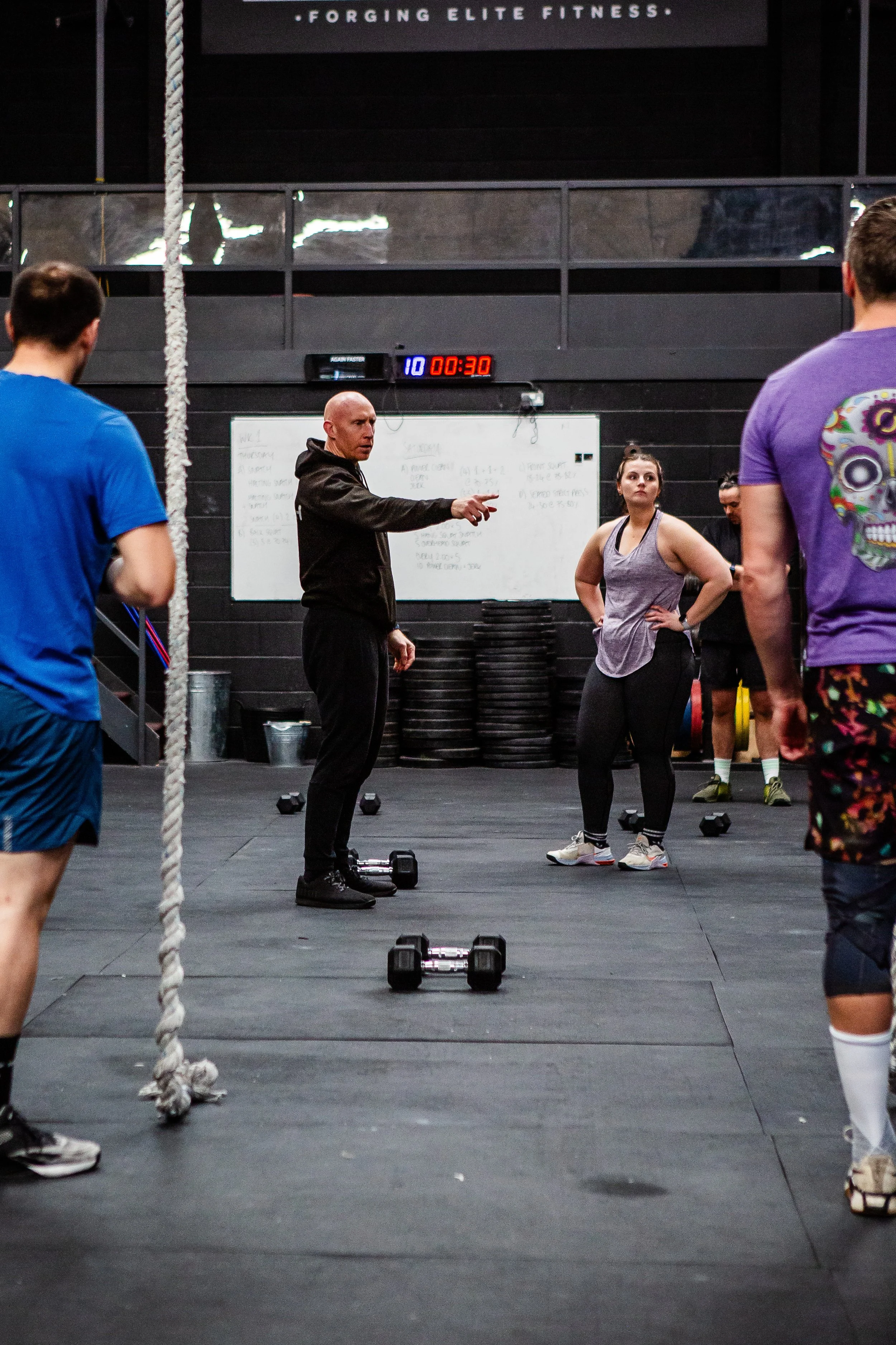 A fitness instructor is giving instructions to a group of people in a CrossFit gym, with exercise equipment like dumbbells on the floor and weight plates stacked in the background. There is a digital timer on the wall showing 30 seconds.