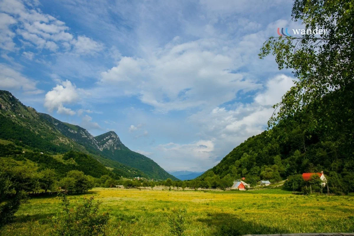 A scenic landscape of a lush green valley with mountains in the background. The sky is blue with scattered clouds, and there are small houses with red roofs on the right side of the valley.