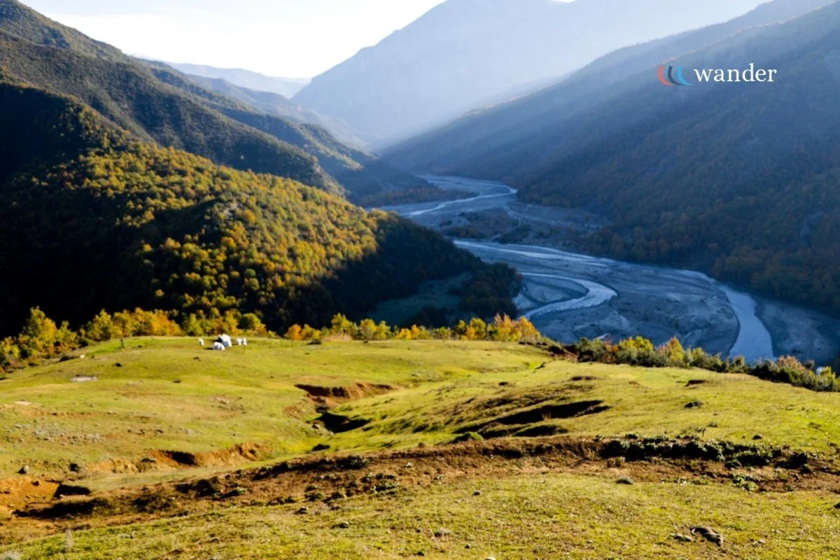 A scenic landscape of a green, grassy field with a few grazing animals, overlooking a river winding through a valley surrounded by forested hills and mountains.