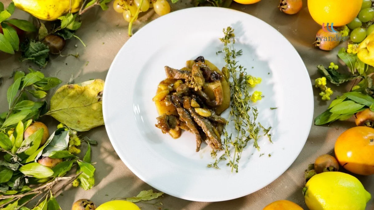 A white plate with a portion of cooked eggplant and vegetables garnished with a sprig of thyme, surrounded by various fruits and leafy herbs.