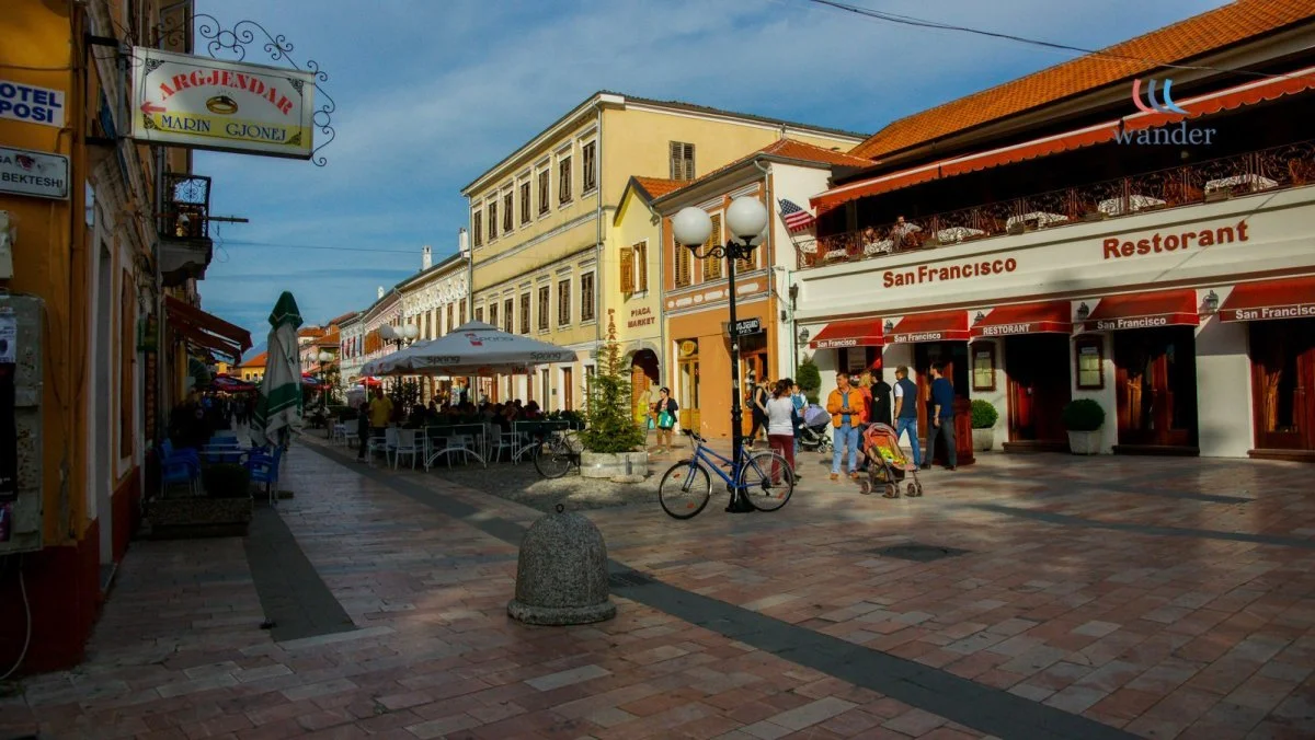 A lively street scene with outdoor café seating, people walking, and historic buildings in a sunny city, likely in San Francisco.