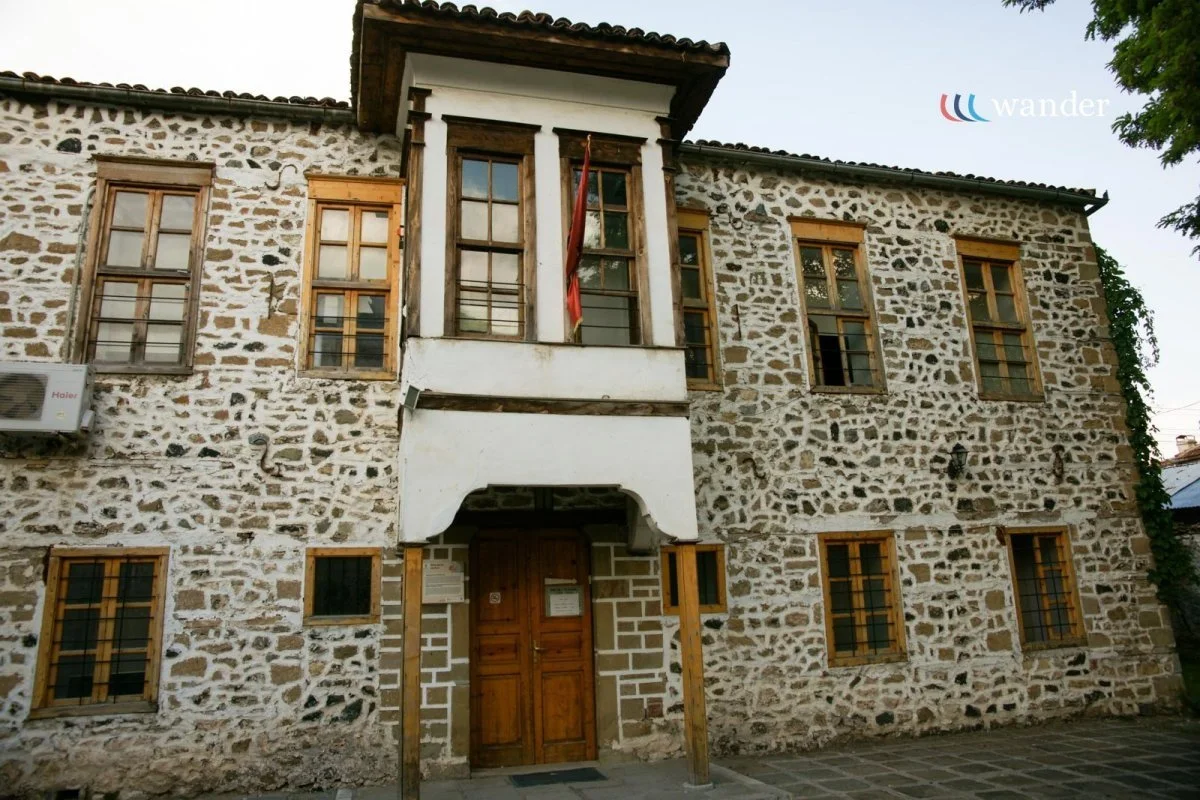 A historic stone building with multiple wooden-framed windows and a wooden door, displaying traditional architecture. There is a sign near the door, an air conditioning unit on the left, and a flag hanging from a balcony.