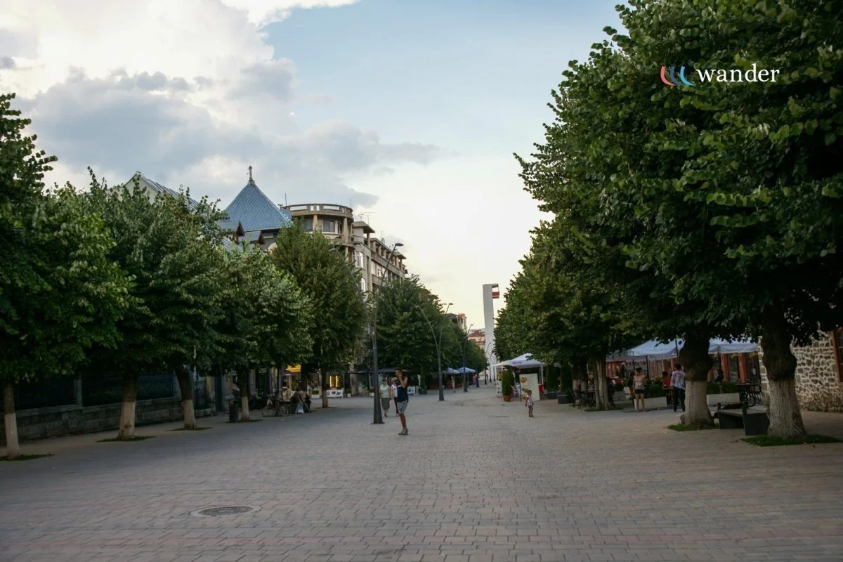 A pedestrian street lined with lush green trees, with a few people walking and cycling, under a partly cloudy sky. Buildings and tents are visible in the background.
