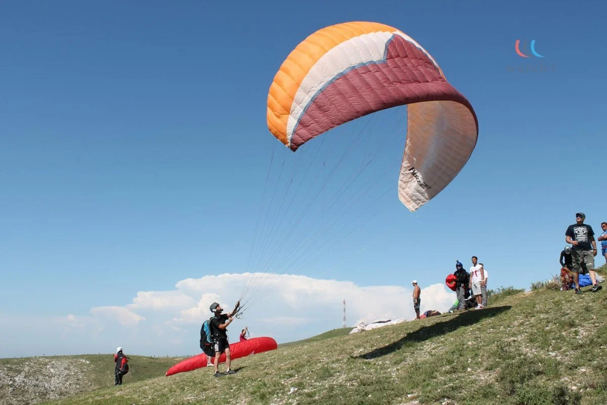 A group of people preparing for paragliding on a grassy hill with a clear blue sky in the background. One person is launching a multicolored parachute into the air.