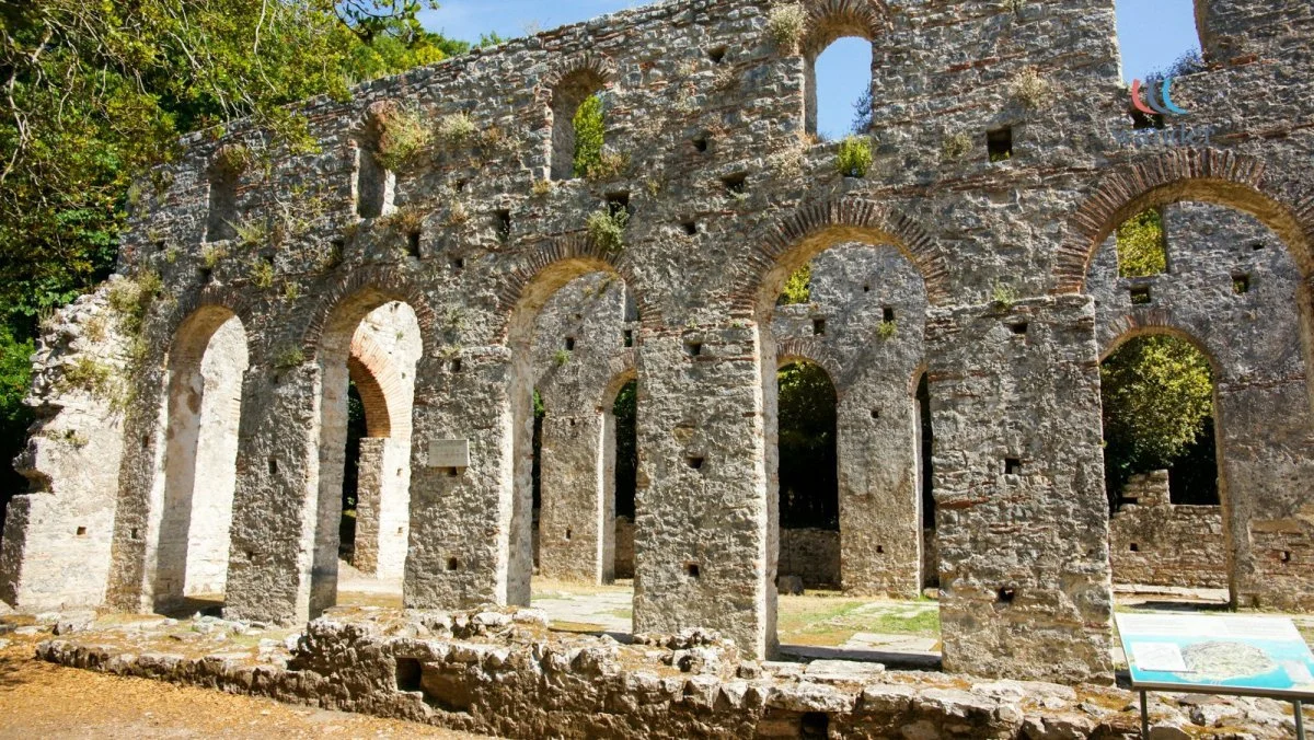 Ruins of an ancient stone structure with large arched openings, surrounded by trees and under a clear blue sky.