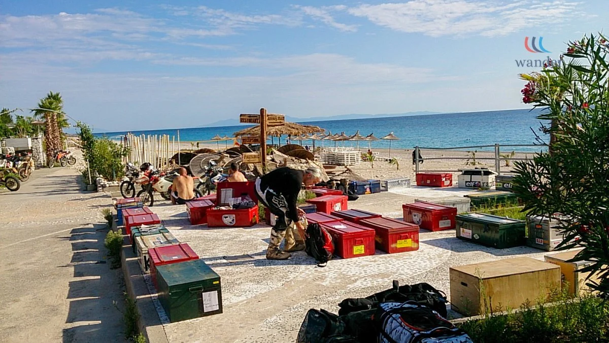 A beachside scene with several large red and green containers on the ground, some people preparing gear, beach umbrellas, and the ocean in the background.