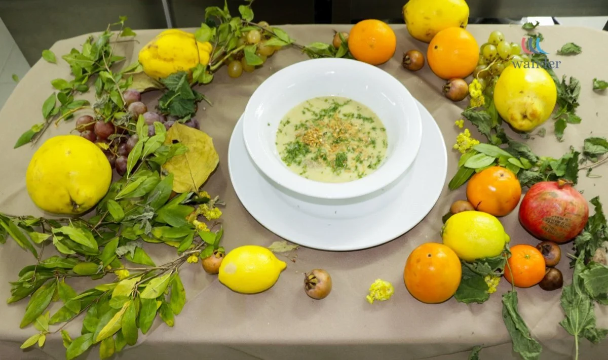 A white bowl of soup garnished with herbs, placed on a plate, surrounded by various fruits such as apples, lemons, oranges, grapes, and small fruits, along with green leaves and vines scattered on a beige table.