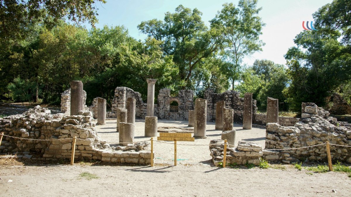 Ancient ruins with stone columns and walls, surrounded by trees on a sunny day.