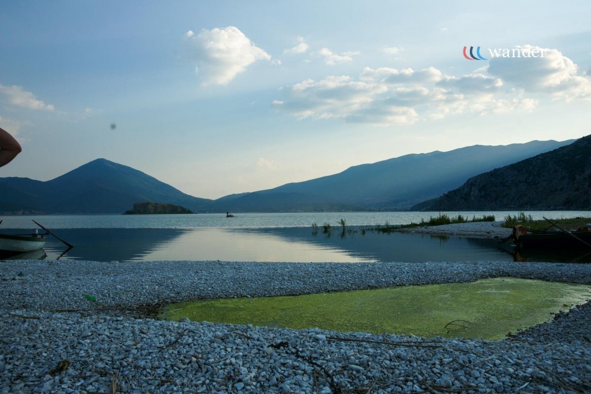 A serene lakeside scene with a pebble beach, calm water, distant mountains, and partly cloudy sky; boats are on the shore with one visible on the left and another on the right.