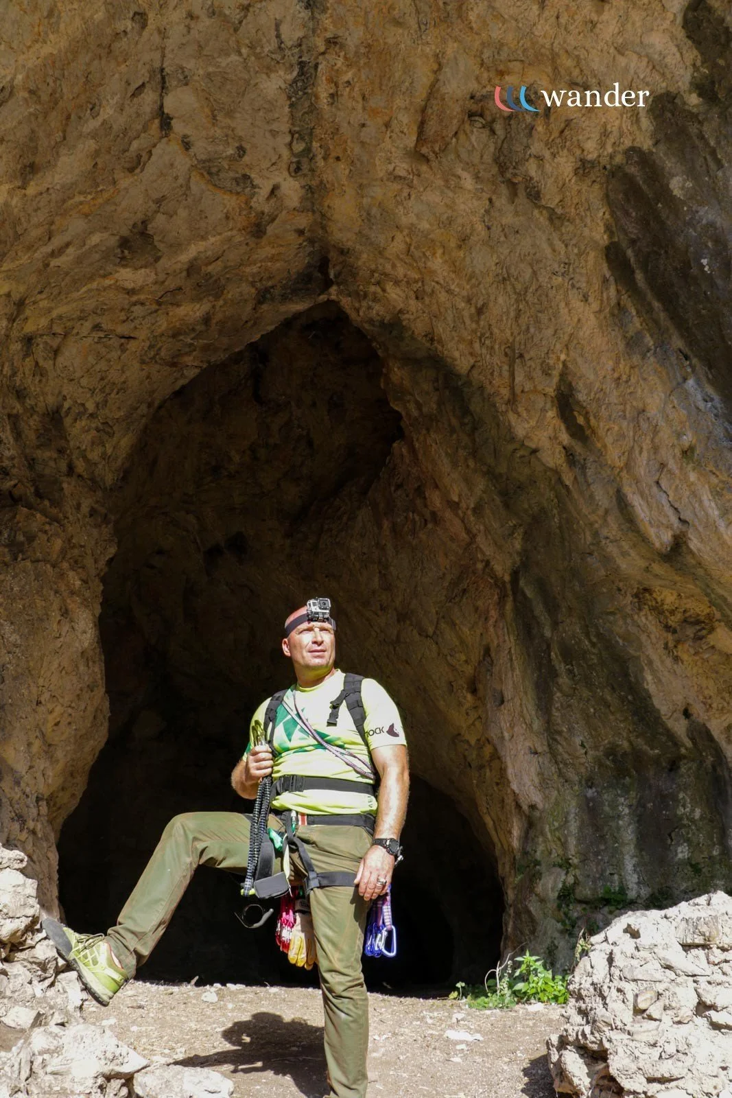 A man standing near the entrance of a cave, wearing outdoor gear, with a GoPro mounted on his head and climbing equipment hanging from his harness.
