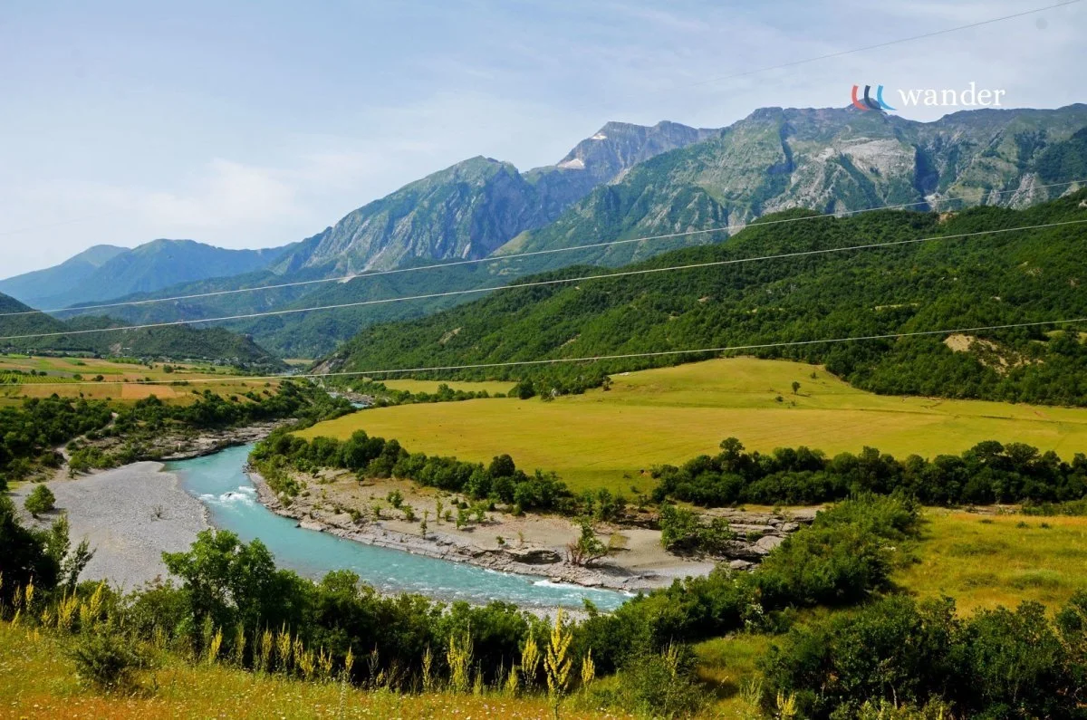 A scenic landscape of rolling green hills, a winding river, and mountains in the background under a partly cloudy sky.