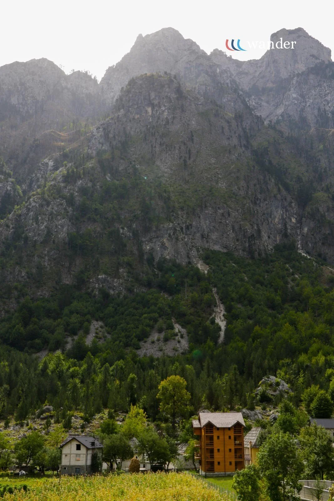 Scenic view of a mountain with lush green trees and a small village at the base, featuring a few houses and a multi-story wooden building.