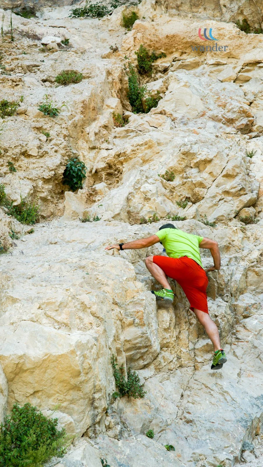A person climbing a steep, rocky outdoor terrain wearing a bright green t-shirt and red shorts with green and black climbing shoes.