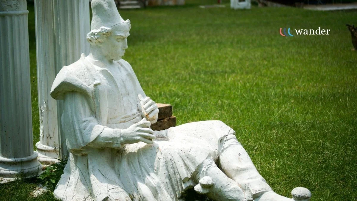 White marble statue of a seated woman wearing a Phrygian cap, holding a small object, set on grass with a column and a wooden block nearby, and the word 'wander' in the top right corner.