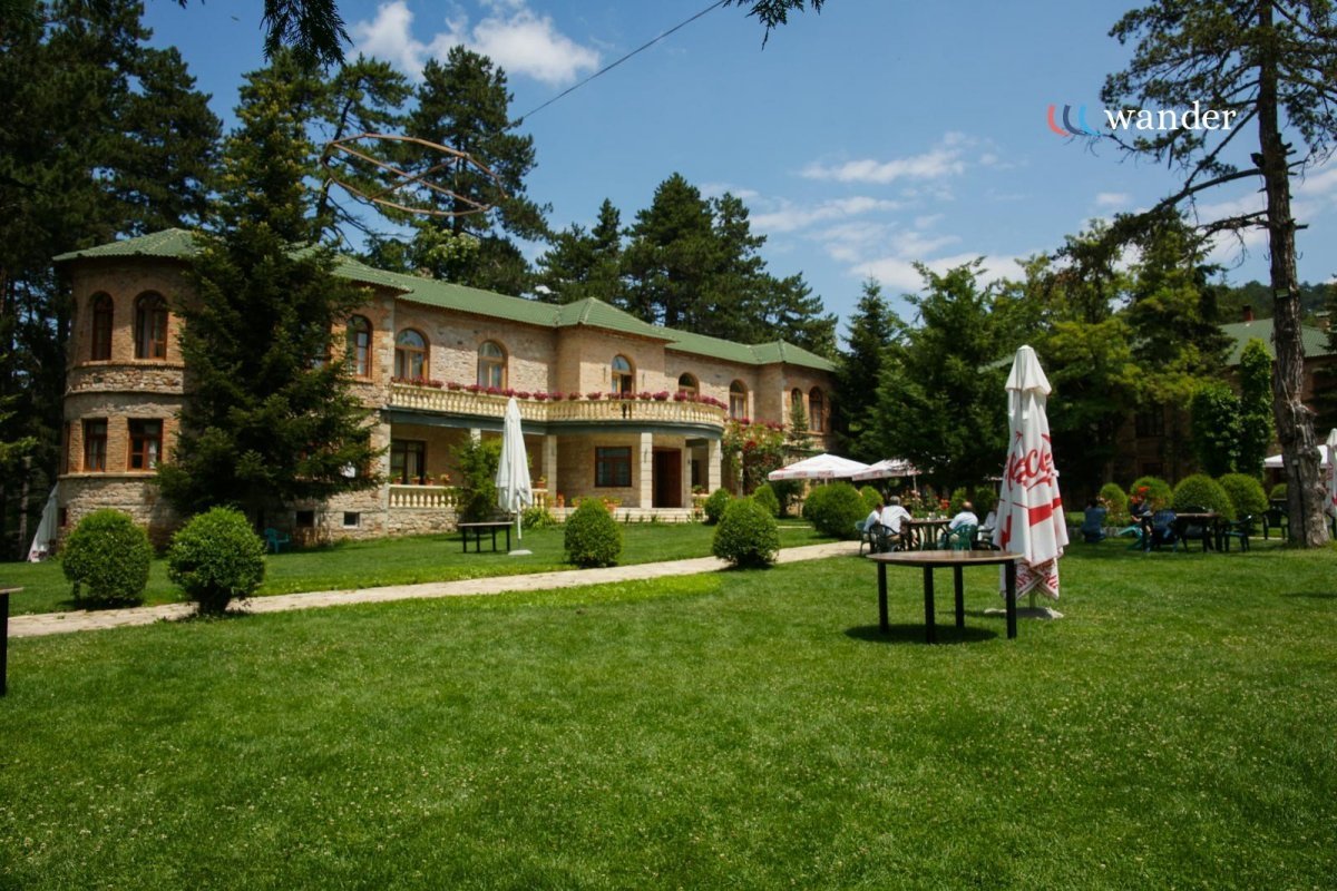 A large stone house with green roof surrounded by well-maintained grass and trees. There are outdoor tables with umbrellas and people sitting, with a clear sky overhead.