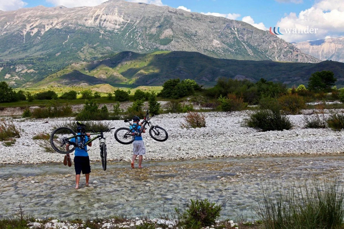 Two children carrying bikes walk through a shallow river with a mountainous landscape in the background.