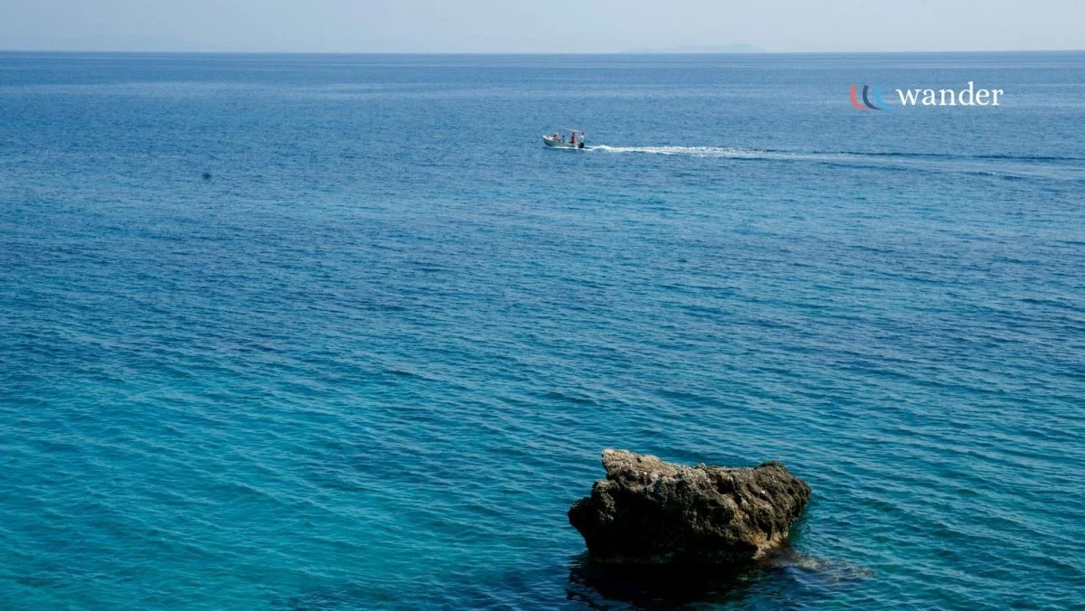 A boat floating on a calm blue ocean with a single rock in the foreground and the word "wander" in the top right corner.