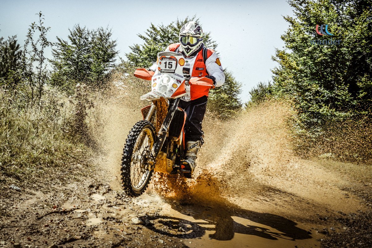 A person riding a dirt bike on an off-road trail during the daytime, kicking up dust and gravel as they jump over a small puddle, surrounded by green trees and bushes.