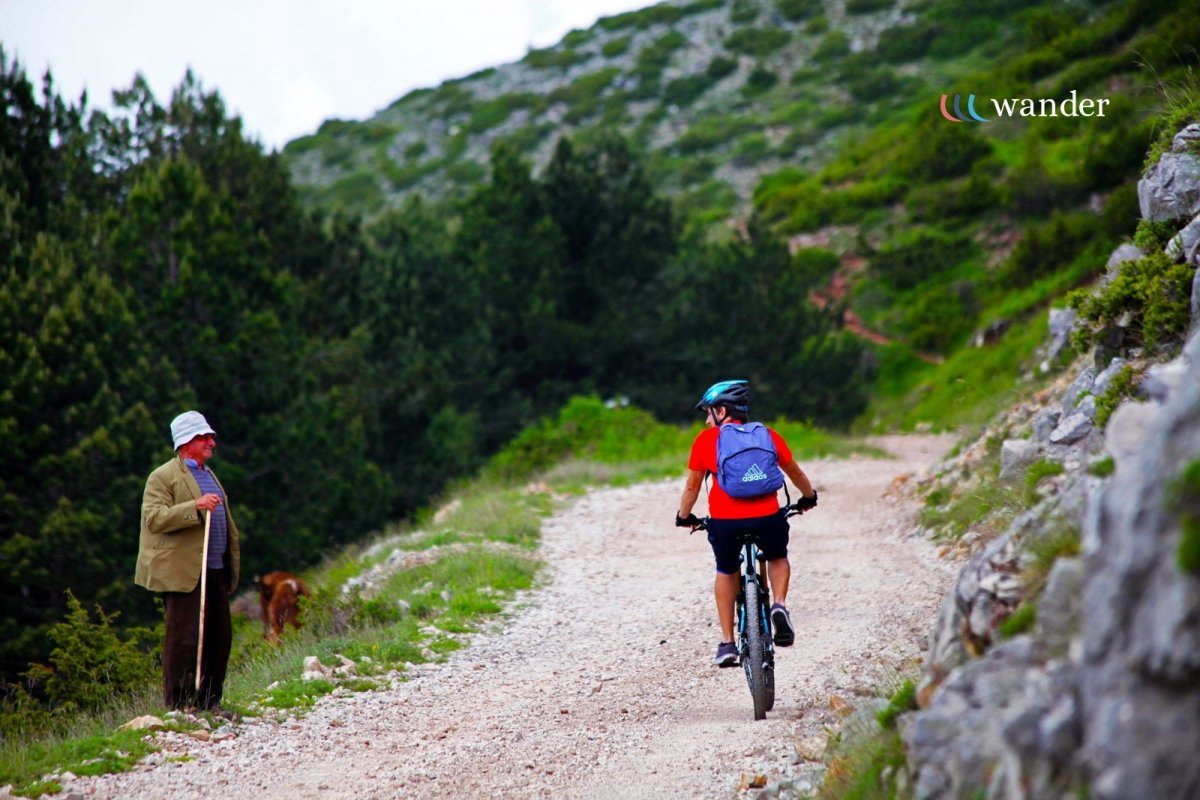 A young boy wearing a red shirt and black shorts riding a mountain bike on a dirt trail in a lush, green mountainous area, with an older woman standing nearby and a dog in the background.