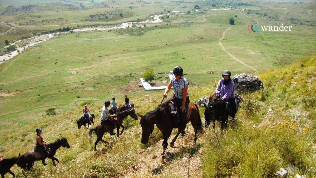 A group of people riding horses uphill through a grassy landscape with hills and a river in the background.