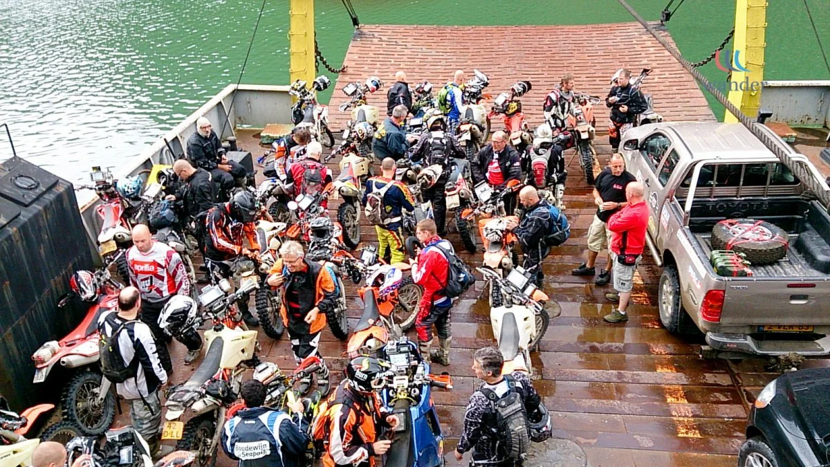 A group of motorcyclists with helmets and gear gather on a ferry dock with motorcycles and a pickup truck, preparing to board a ferry crossing a body of water.