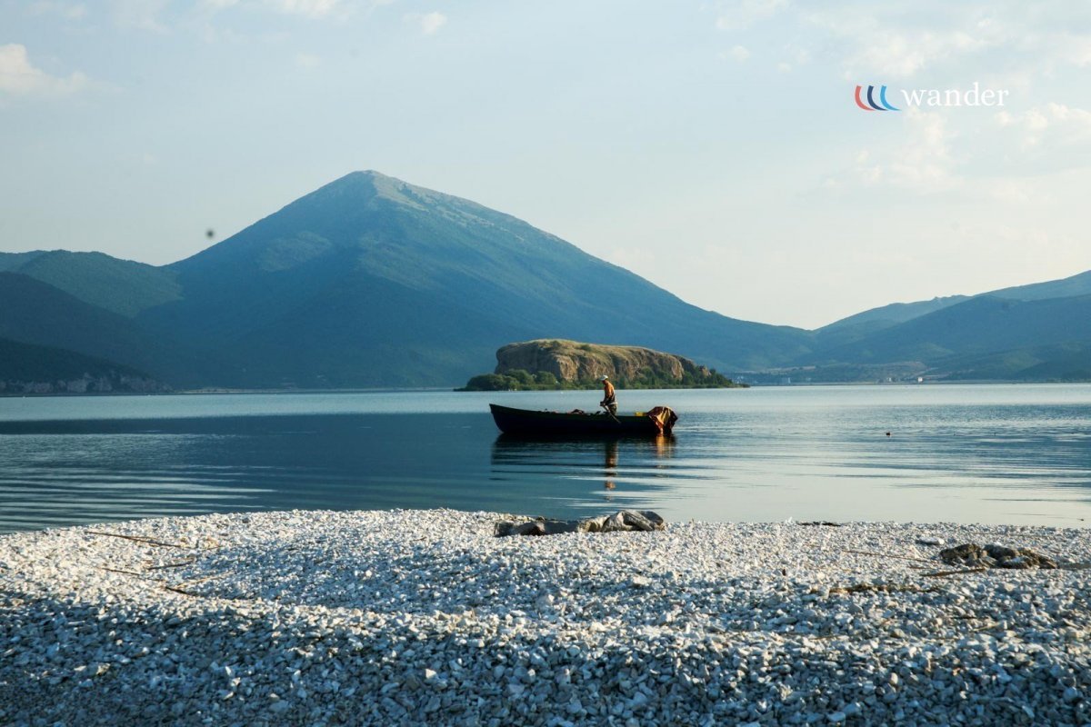 Person in a small boat on a calm lake with mountains in the background and a pebble beach in the foreground.