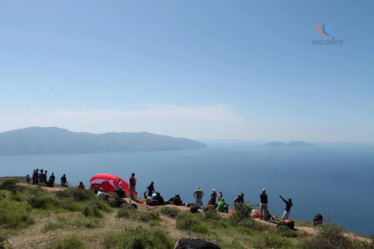 Group of people watching and preparing for paragliding on a grassy hilltop overlooking a large body of water and distant mountains under a clear blue sky.