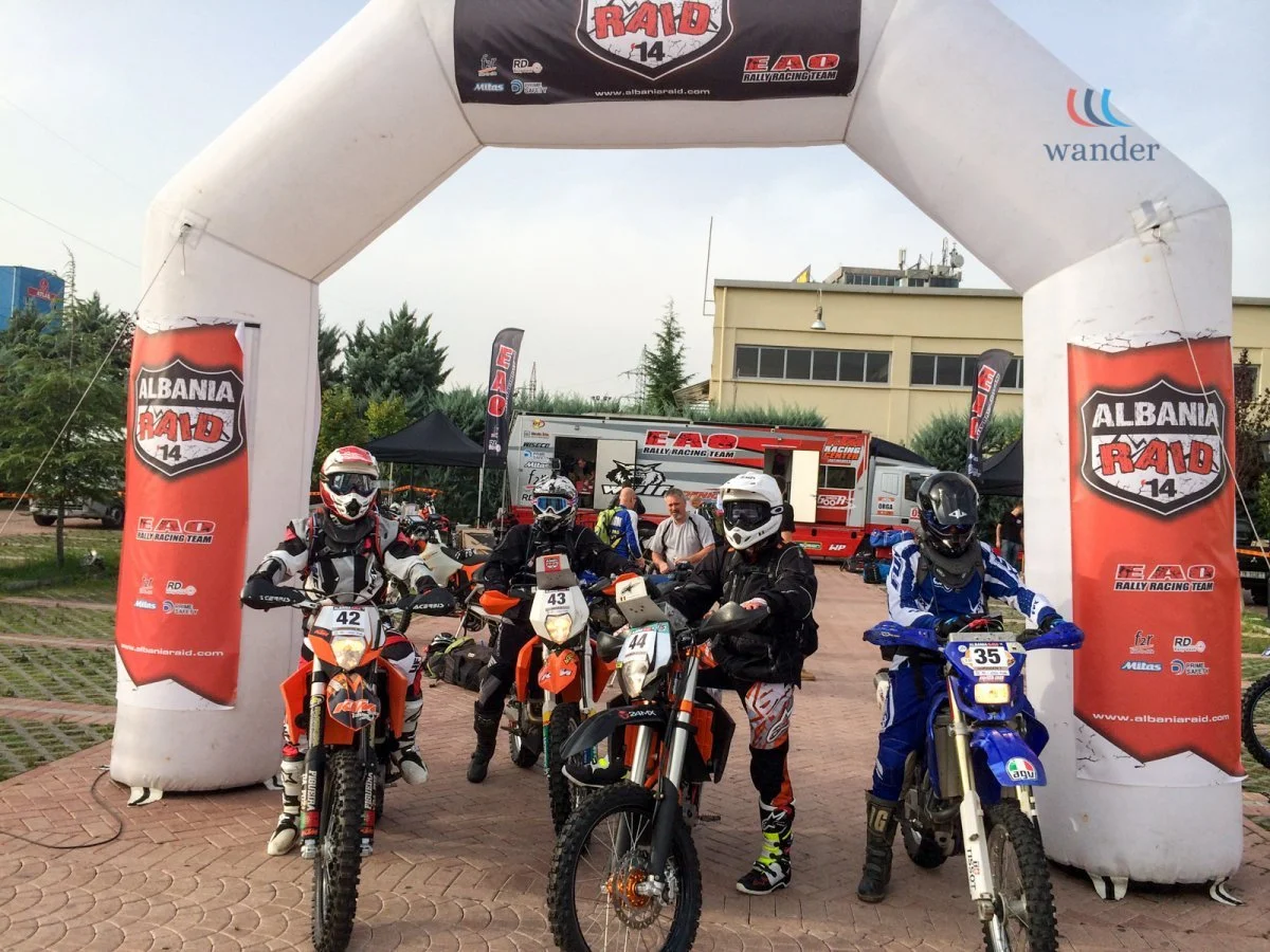 Group of four motorcyclists in full riding gear on KTM dirt bikes at an outdoor rally event under an inflatable archway with 'Albania Raid 14' banners. A mobile event setup with signage and people in the background.
