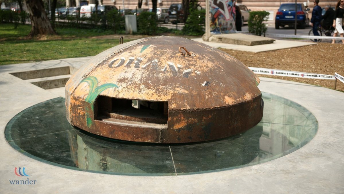 A rusted, dome-shaped object with small rectangular opening, displayed on a circular glass platform outdoors, with graffiti and people visible in the background.