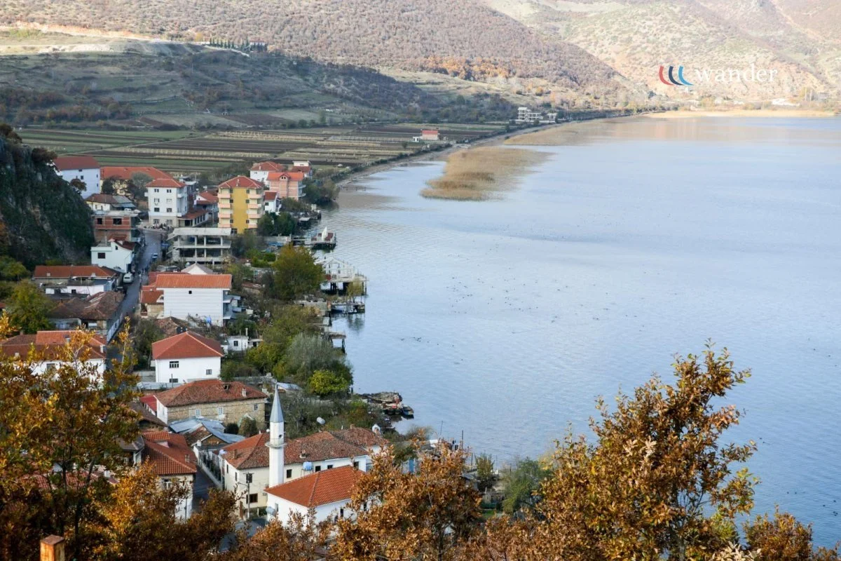 A small lakeside village with houses and a mosque along the shoreline, surrounded by hills and trees with autumn foliage.