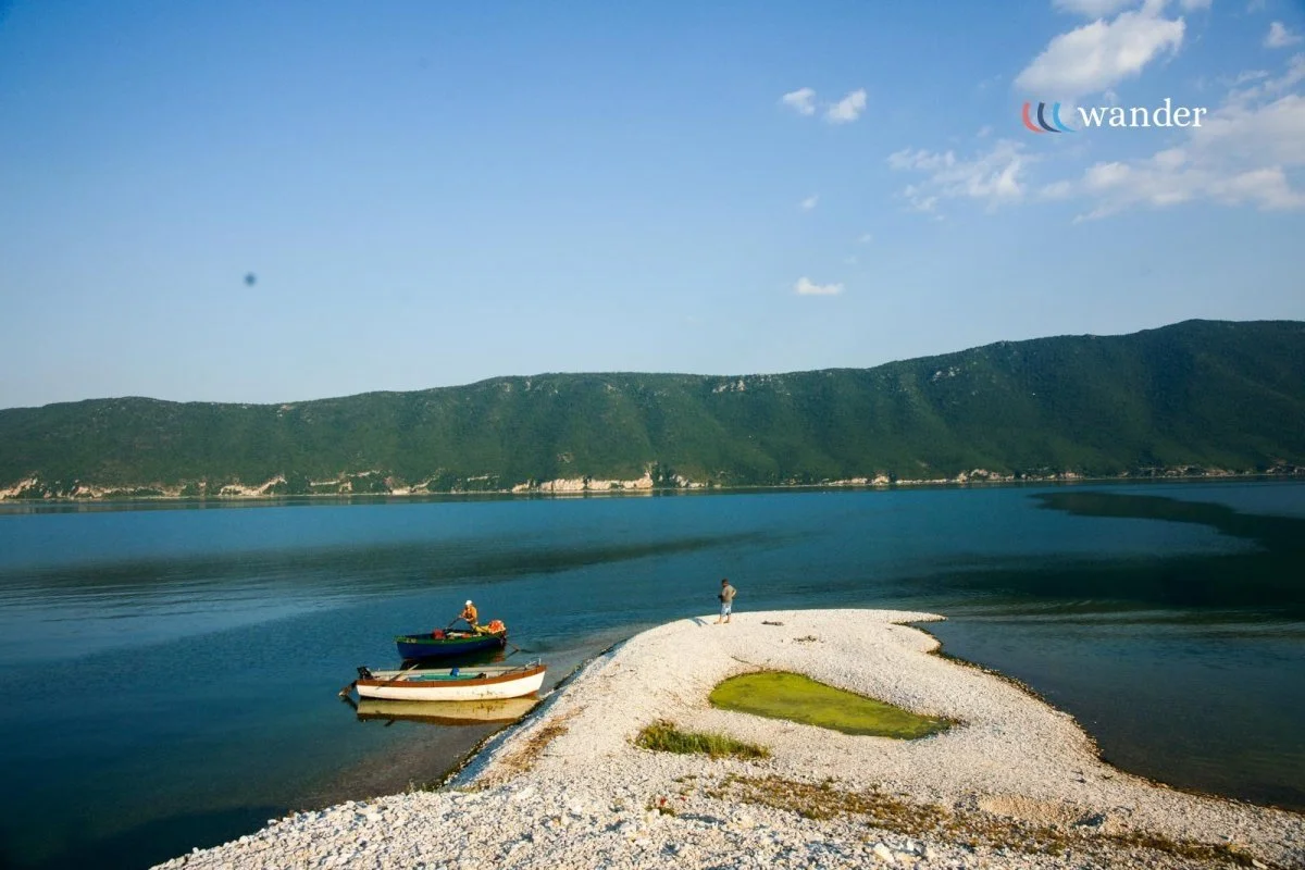A tranquil lakeside scene with two boats docked on a small pebble beach, a person standing on the beach, and a backdrop of green hills under a blue sky with a few clouds.