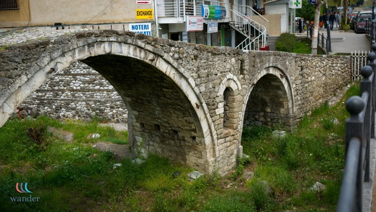 Ancient stone bridge with arches over a grassy area, situated next to modern buildings and a sidewalk with people walking.