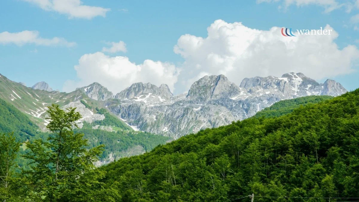A scenic mountain landscape with green trees in the foreground, rugged snow-capped peaks in the background, and a partly cloudy sky.