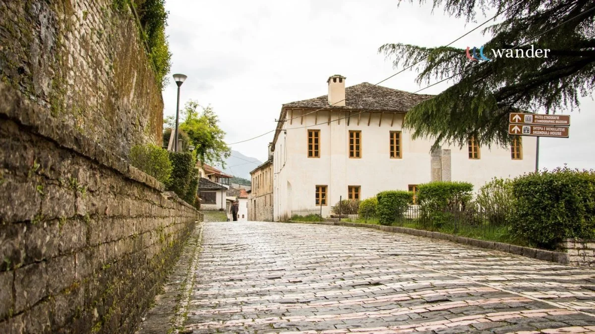 A cobblestone street in a European village with a white building, bushes, and trees, under an overcast sky.