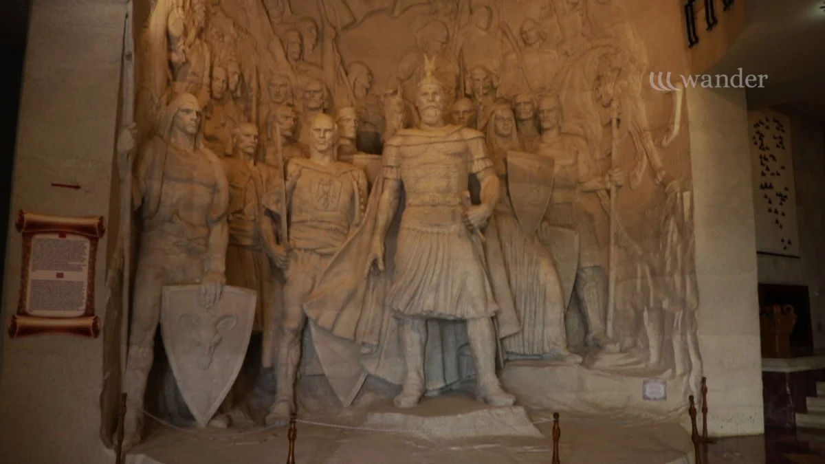 A detailed stone relief sculpture depicting a group of Roman soldiers and a central figure dressed as a Roman emperor or general, holding a staff, with soldiers holding shields and wearing helmets, set in an indoor museum setting.